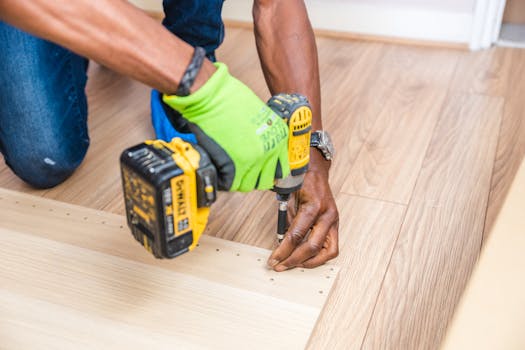 Home Close-up of handyman drilling wood indoors with green gloves and cordless drill.
